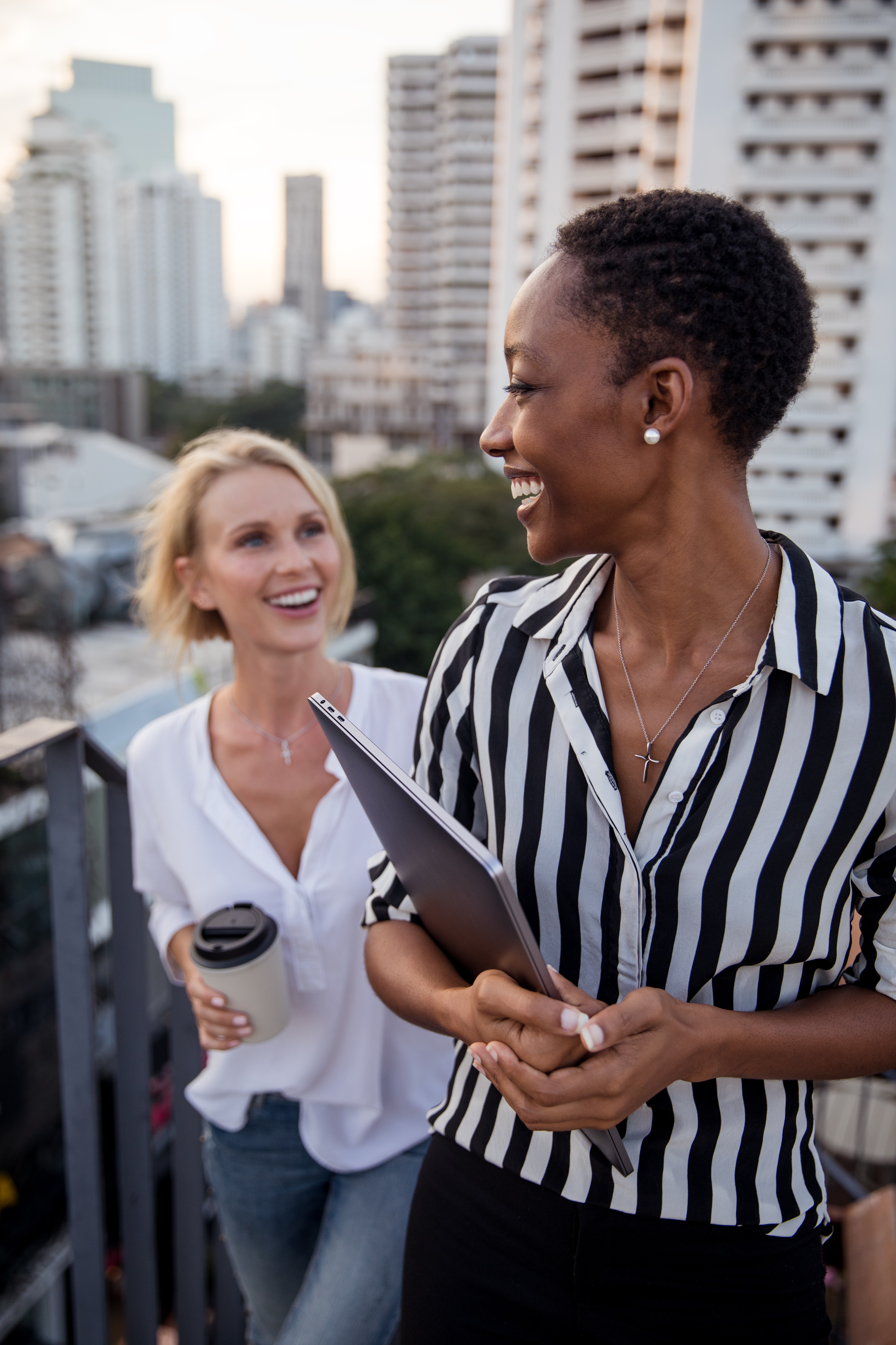 Two women stand outside, each holding a coffee cup in hand, and smiling at each other.