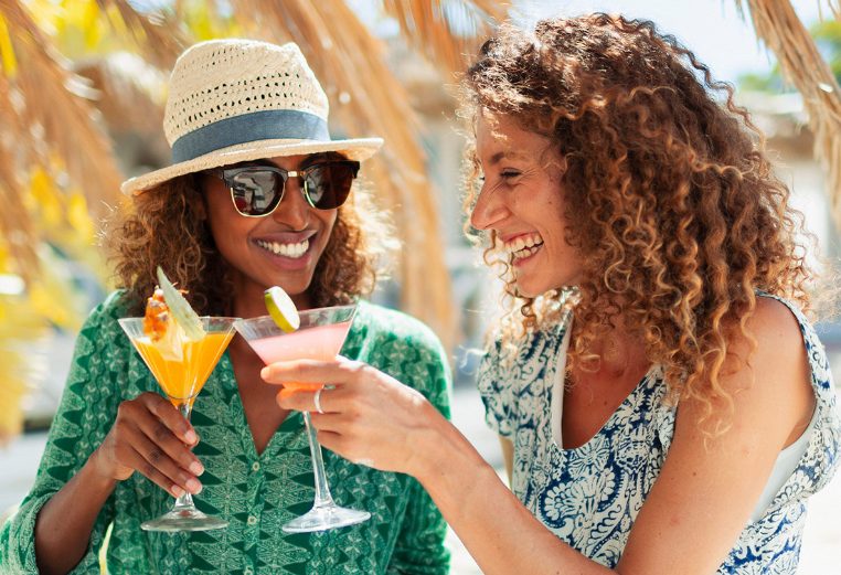 Two women enjoying cocktails and smiling while another woman sits in the background looking at her phone.