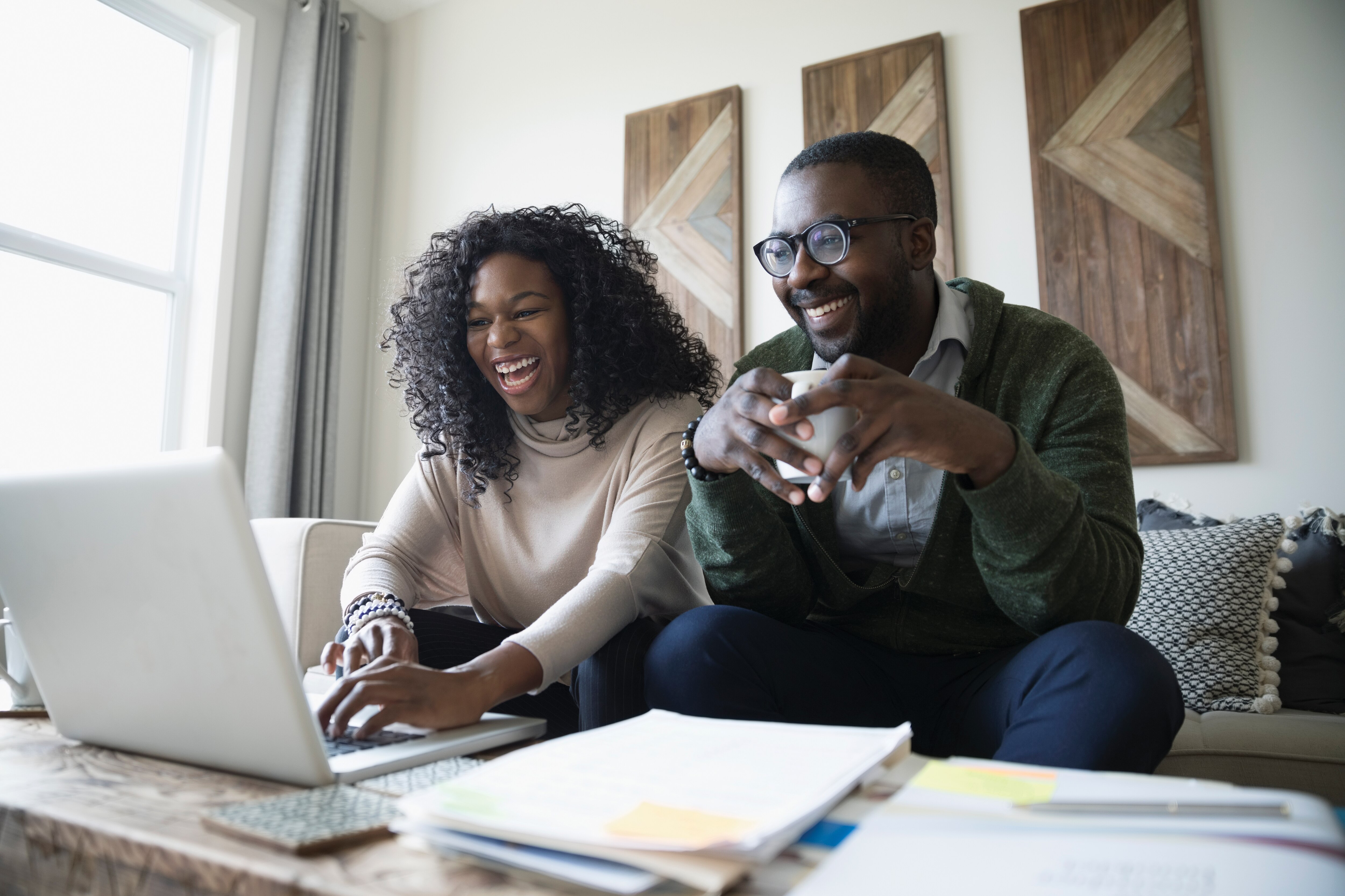 Laughing couple with laptop drinking coffee and paying bills online in living room